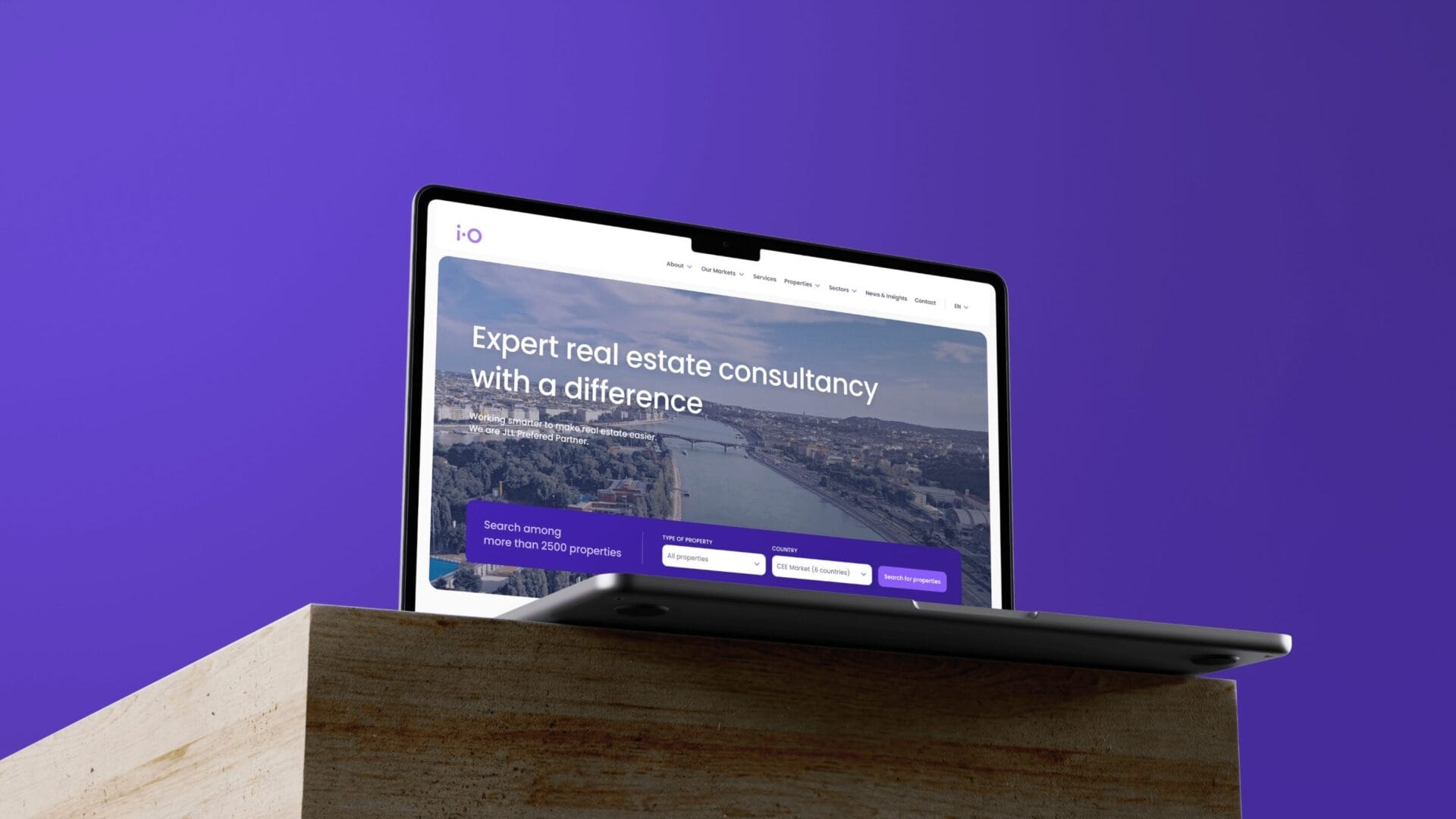 A laptop on violet showing the iO Partners homepage: “Expert real estate consultancy with a difference” above a search bar.