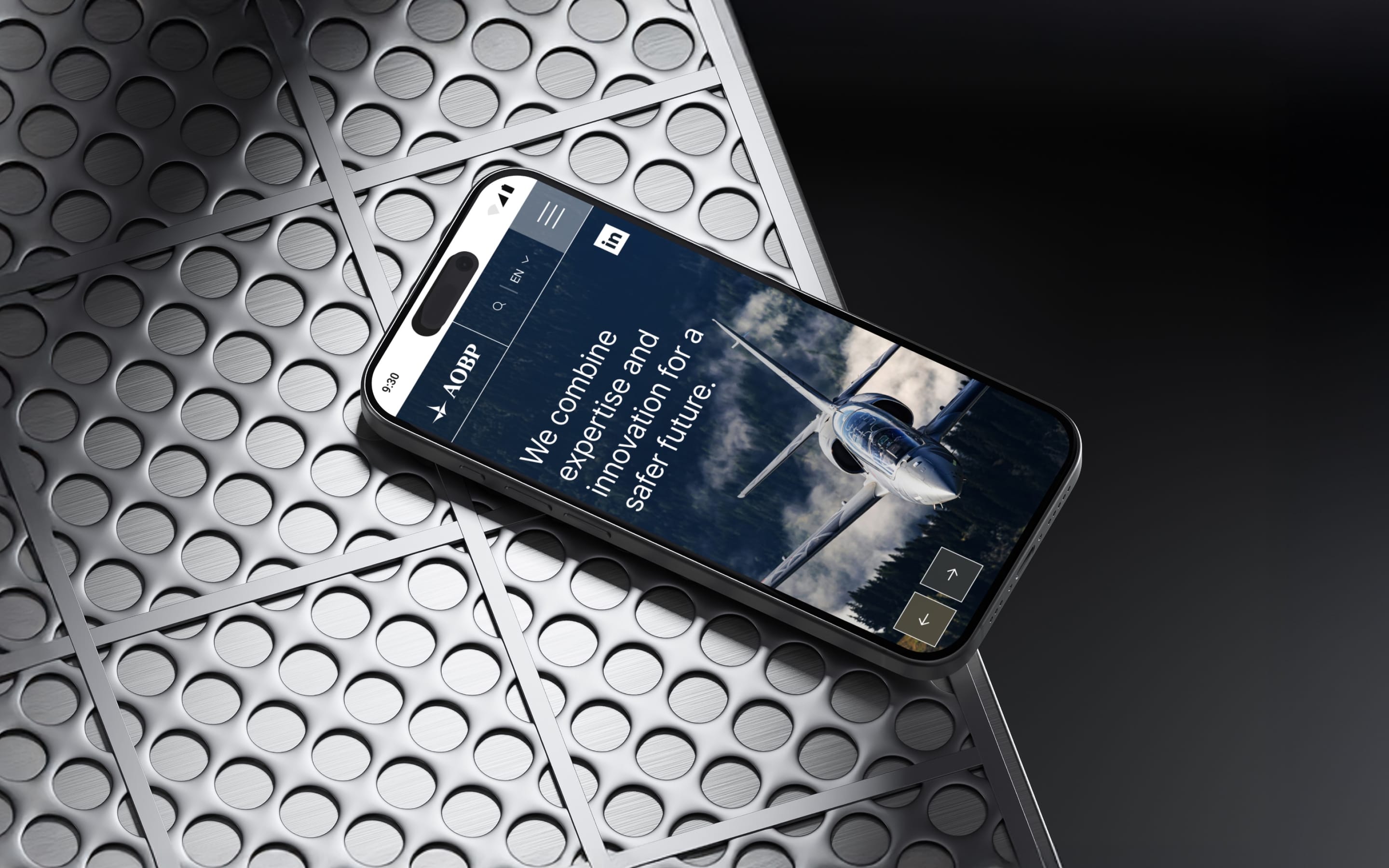 A phone on a perforated steel plate showing the AOBP homepage: “We combine expertise and innovation for a safer future.”