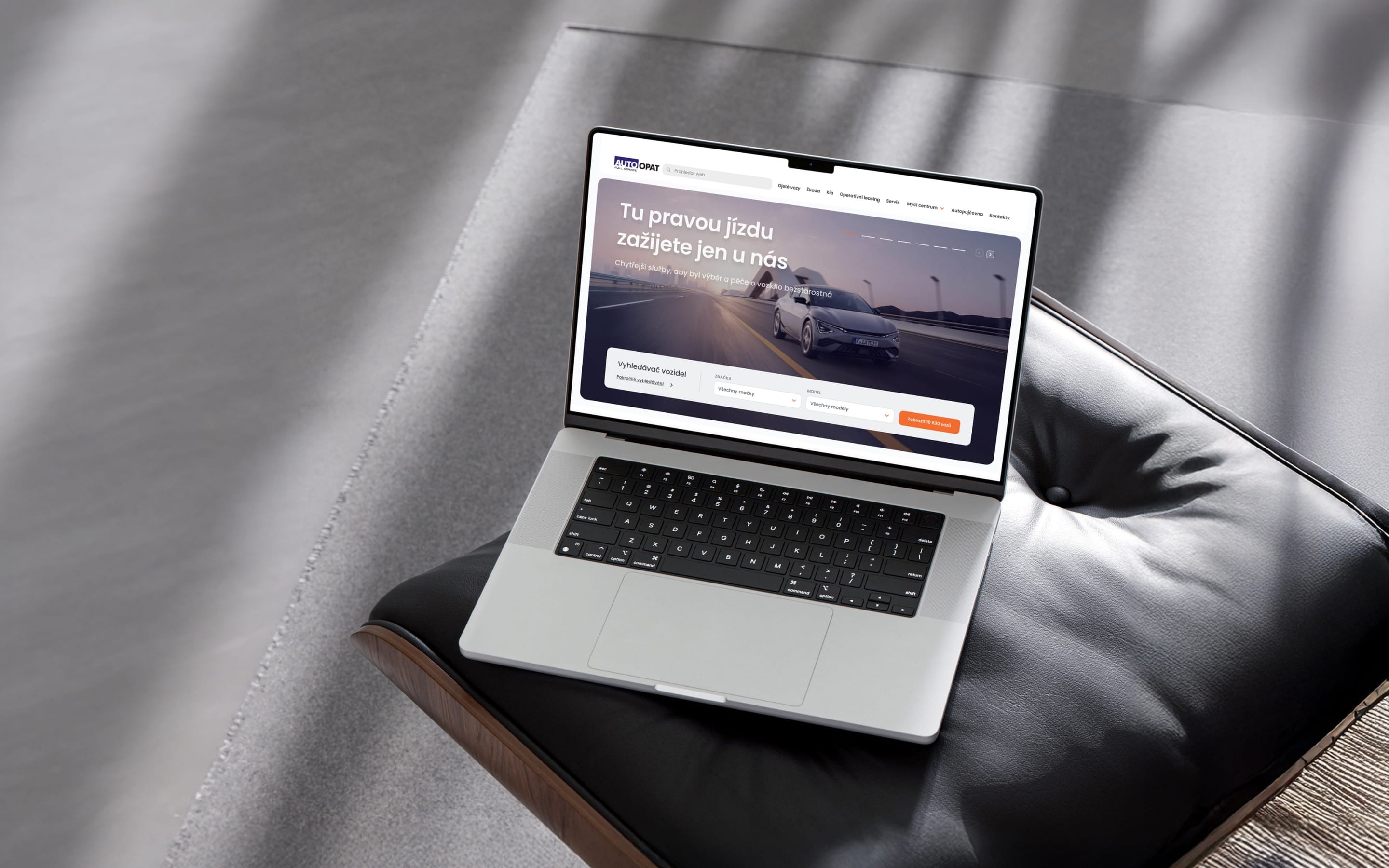 A laptop on a black leather cushion showing the AutoOpat homepage, with a car on a bridge and the vehicle search panel.