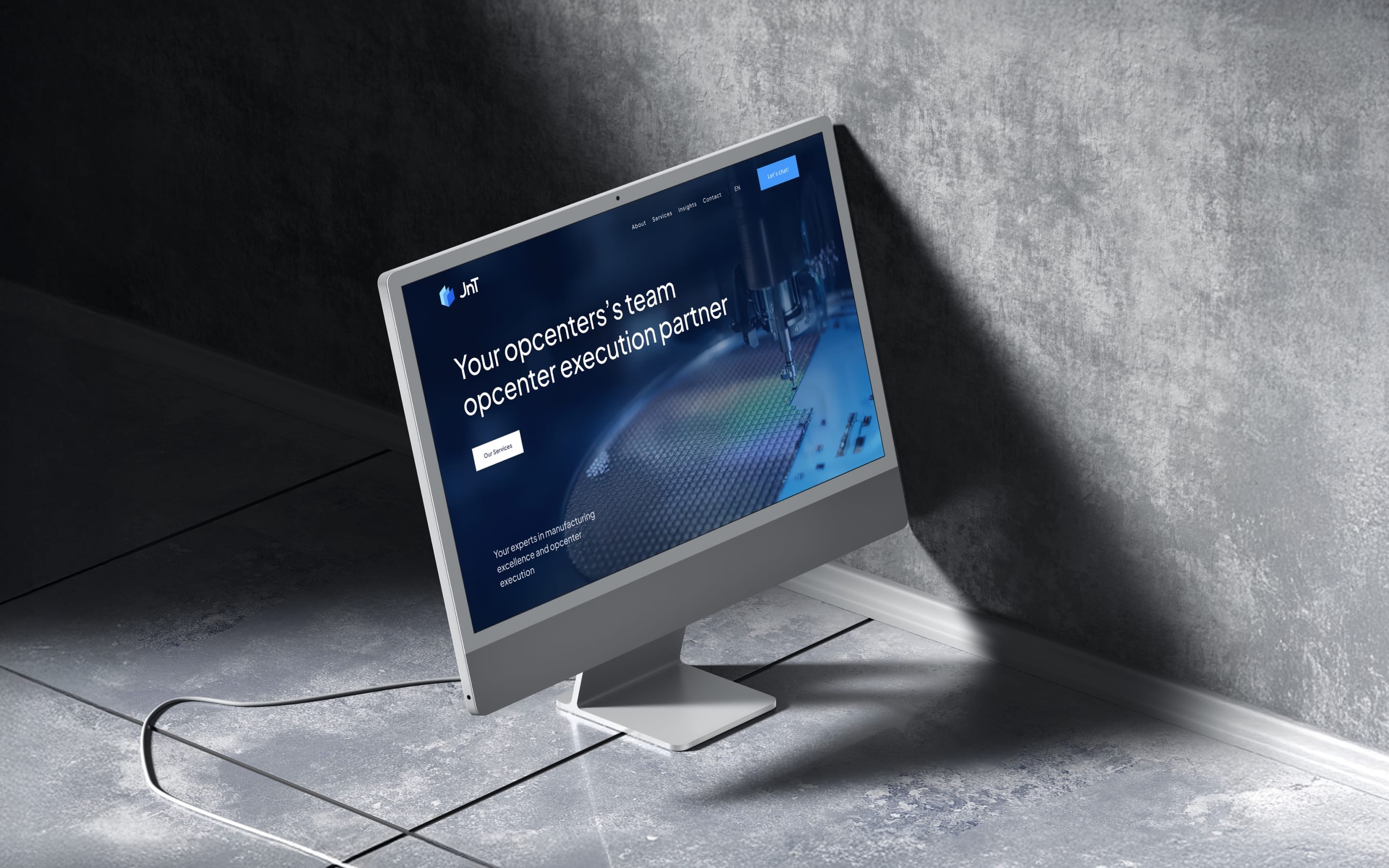 An iMac on a concrete floor in raking light, showing the JNT Digital homepage over a deep blue manufacturing image.