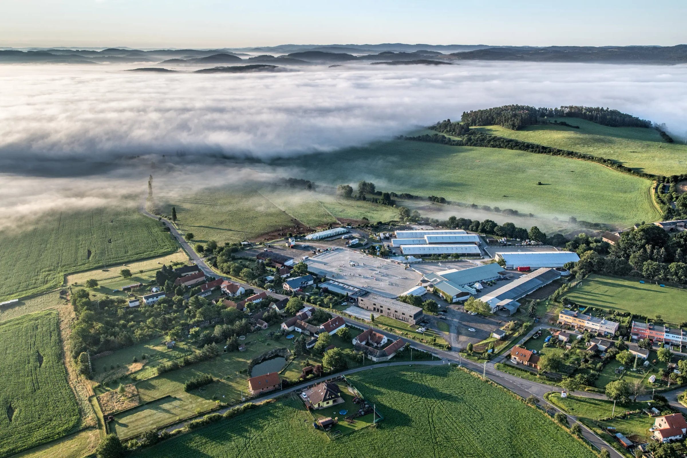 An aerial photo of the equestrian centre at dawn: arenas, stables and halls among green fields, with mist over the hills.