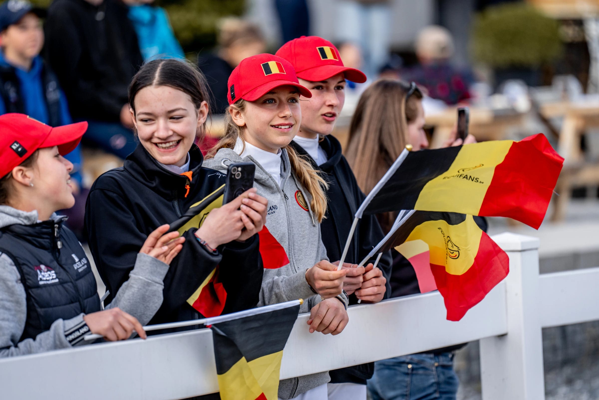 Young fans in red caps wave Belgian flags and film on their phones at the white arena rail.