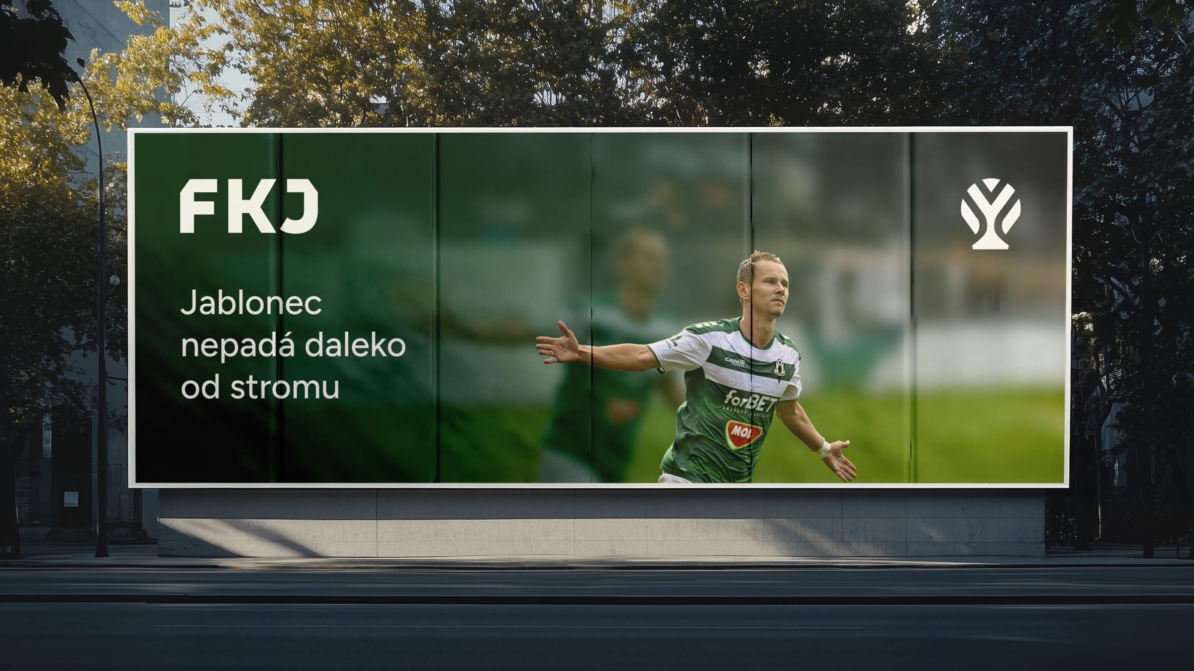 A street billboard with the FKJ wordmark, apple-tree symbol and a celebrating player: “Jablonec nepadá daleko od stromu”.