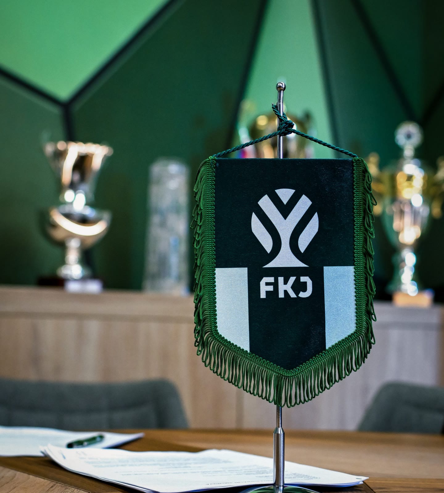 A dark green FKJ pennant with the new symbol on a meeting table, trophies behind it.