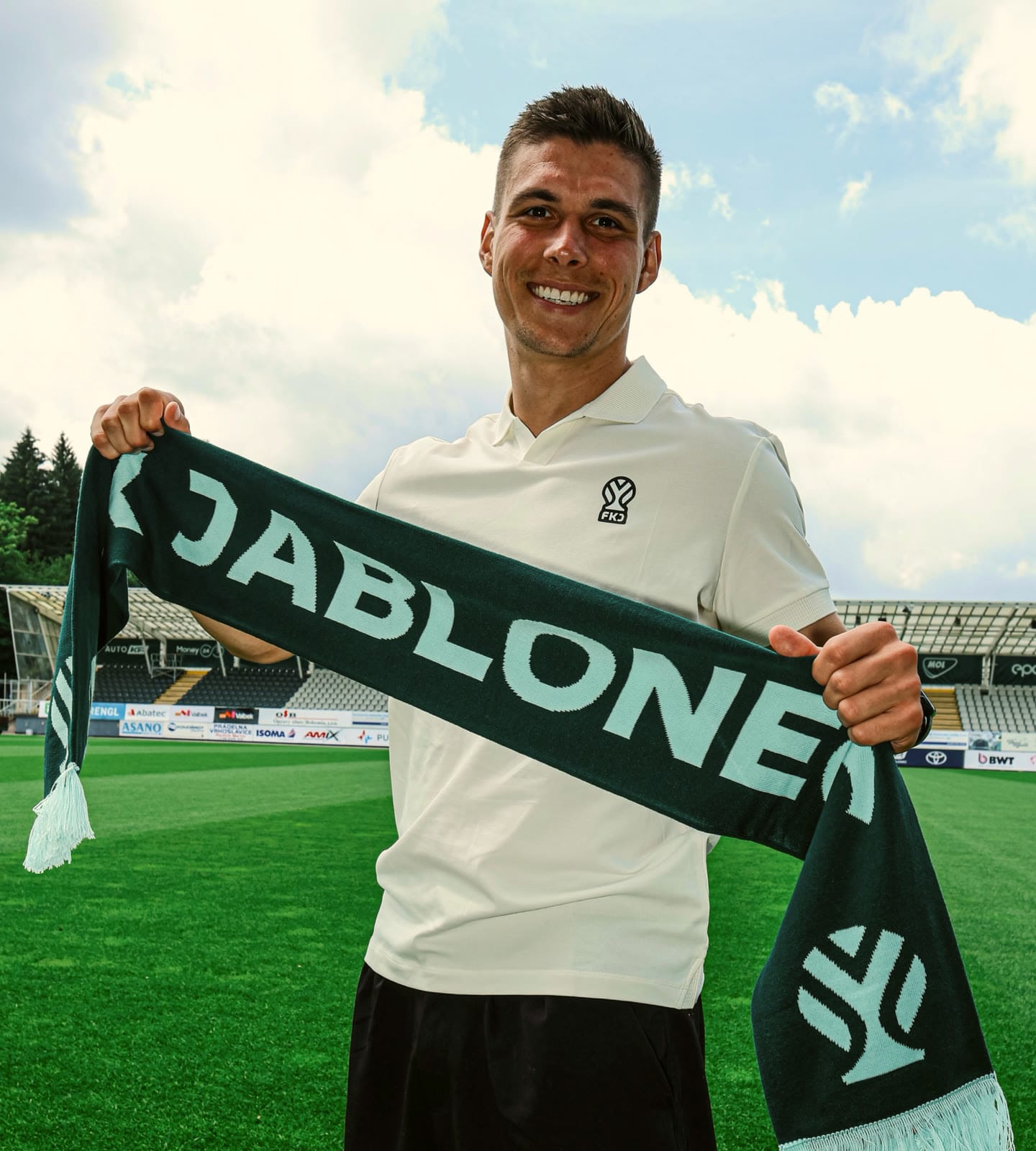 A smiling player on the pitch holding up a dark green Jablonec scarf with the new symbol.