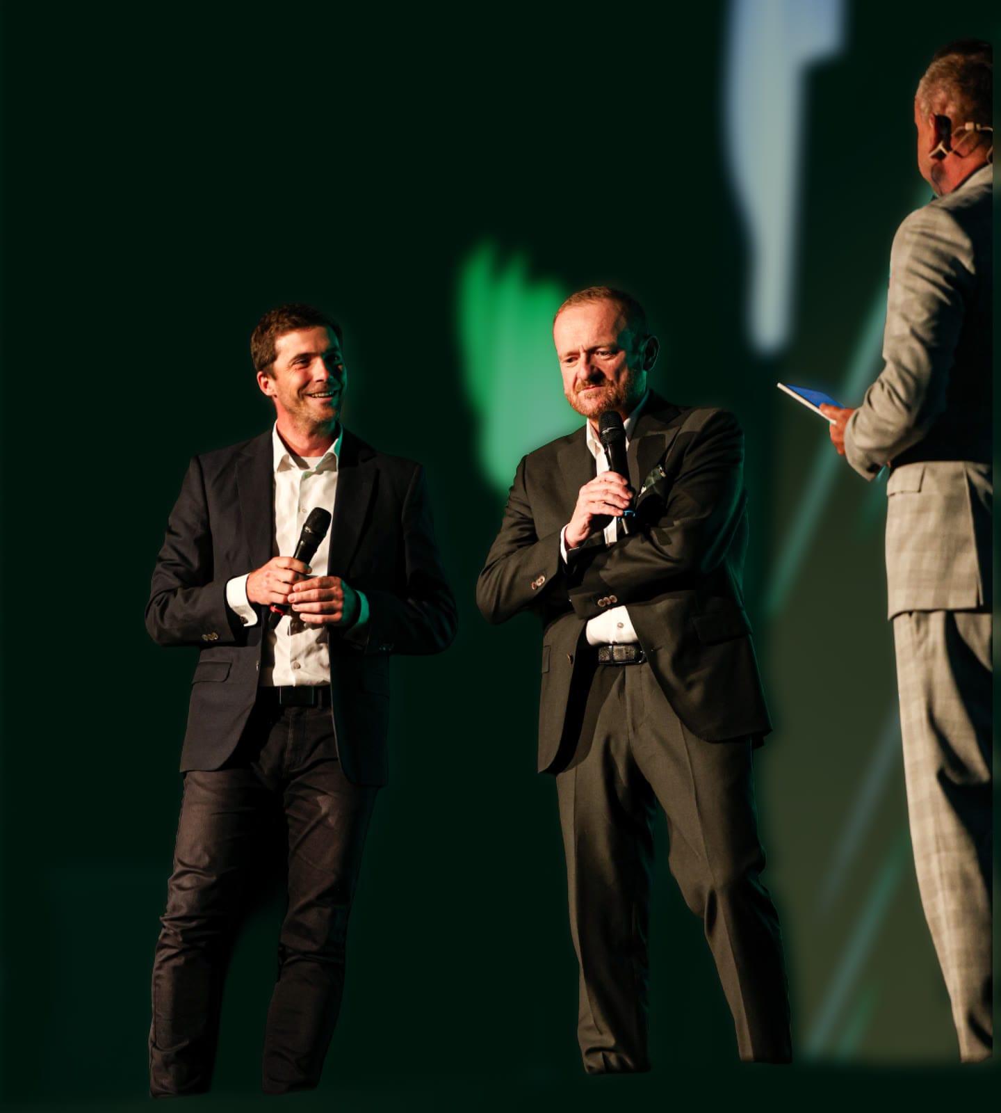 Two guests with microphones on stage at the gala evening, the host beside them.