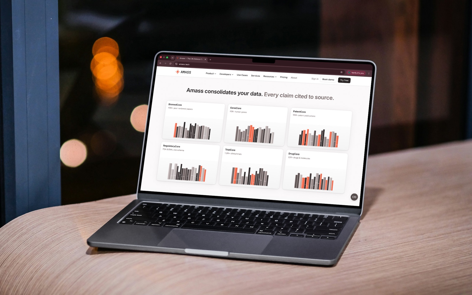 A laptop on a wooden desk showing the Amass homepage: “Amass consolidates your data. Every claim cited to source.”